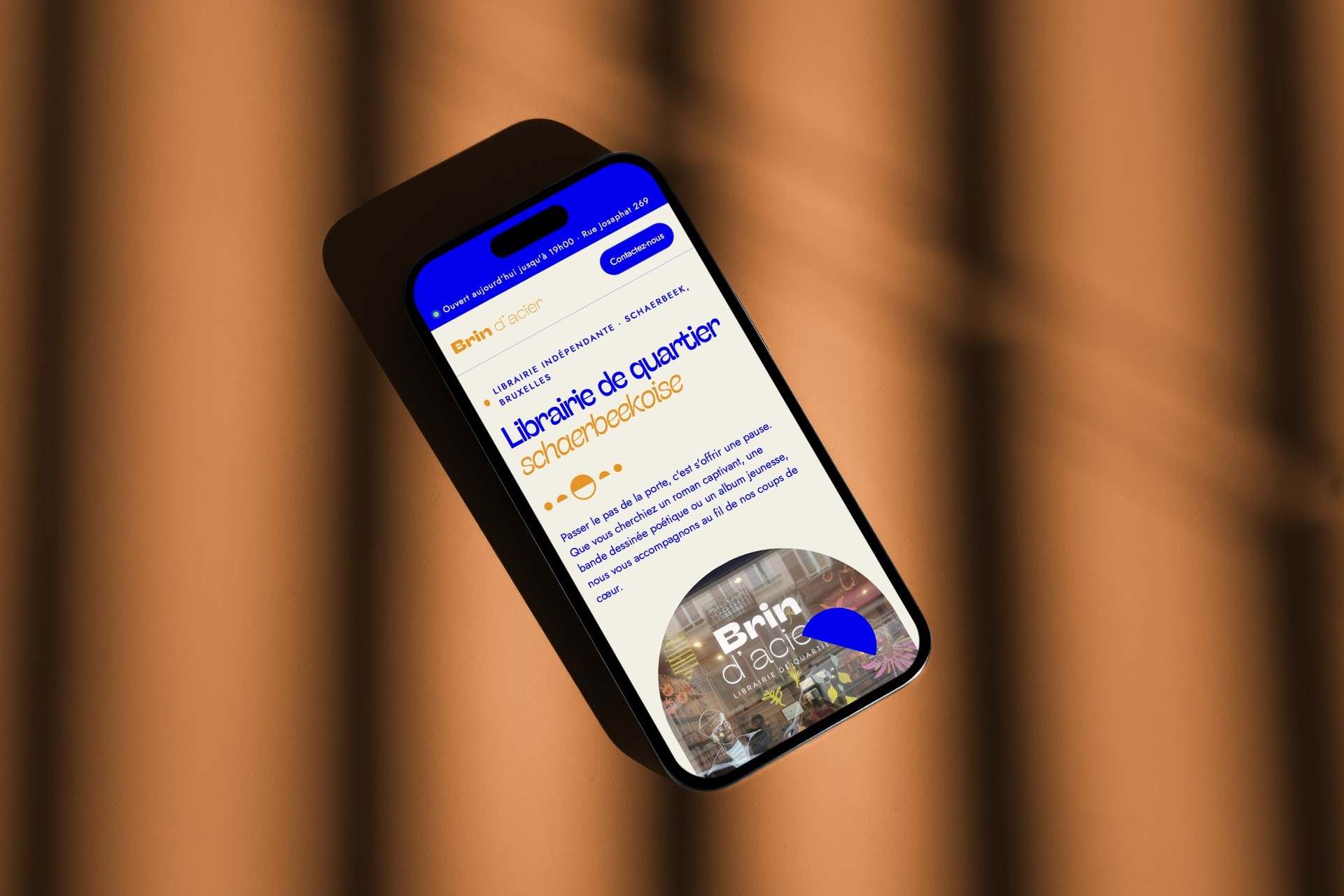 The Brin d'acier site on a phone lying on an orange surface: a neighbourhood bookshop in Schaerbeek, with the shopfront cropped into a half circle.