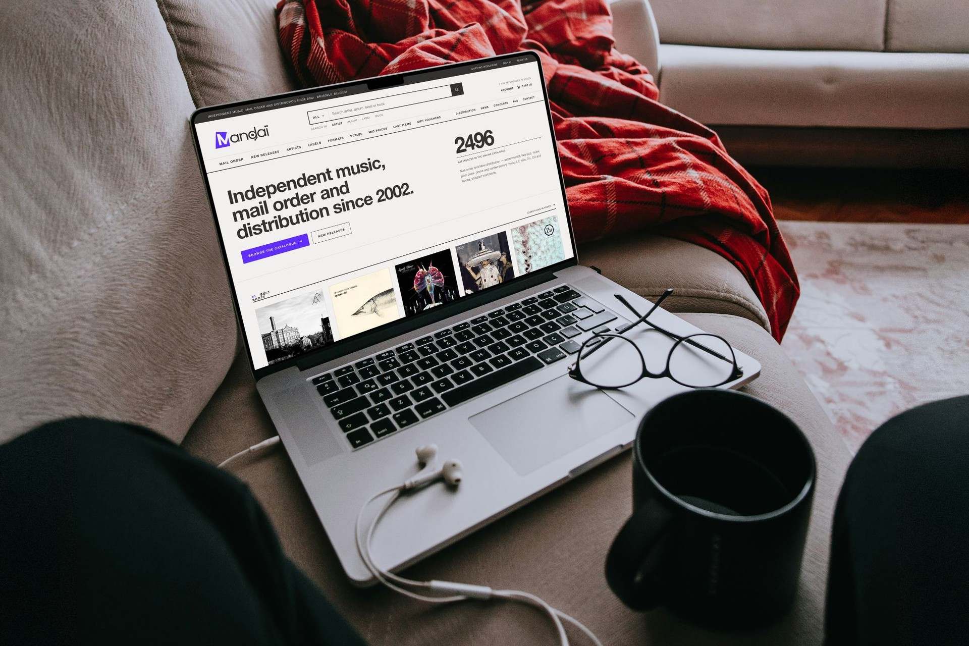 The Mandaï site on a laptop resting on a sofa: independent music, mail order and distribution since 2002, 2,496 references and a row of record covers.