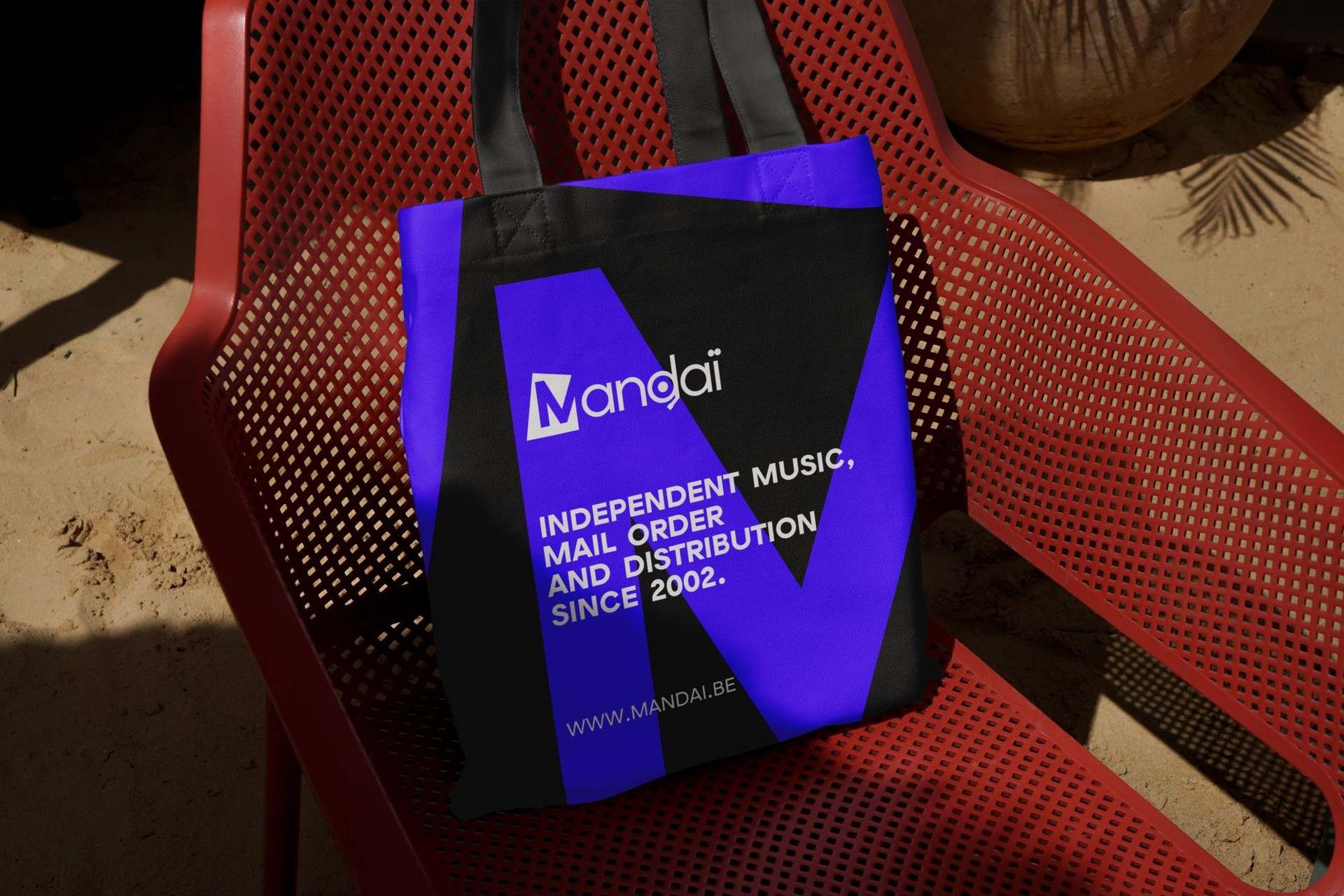 A black and violet Mandaï tote bag on a red perforated chair, a giant M cut across it under the shop's line and web address.
