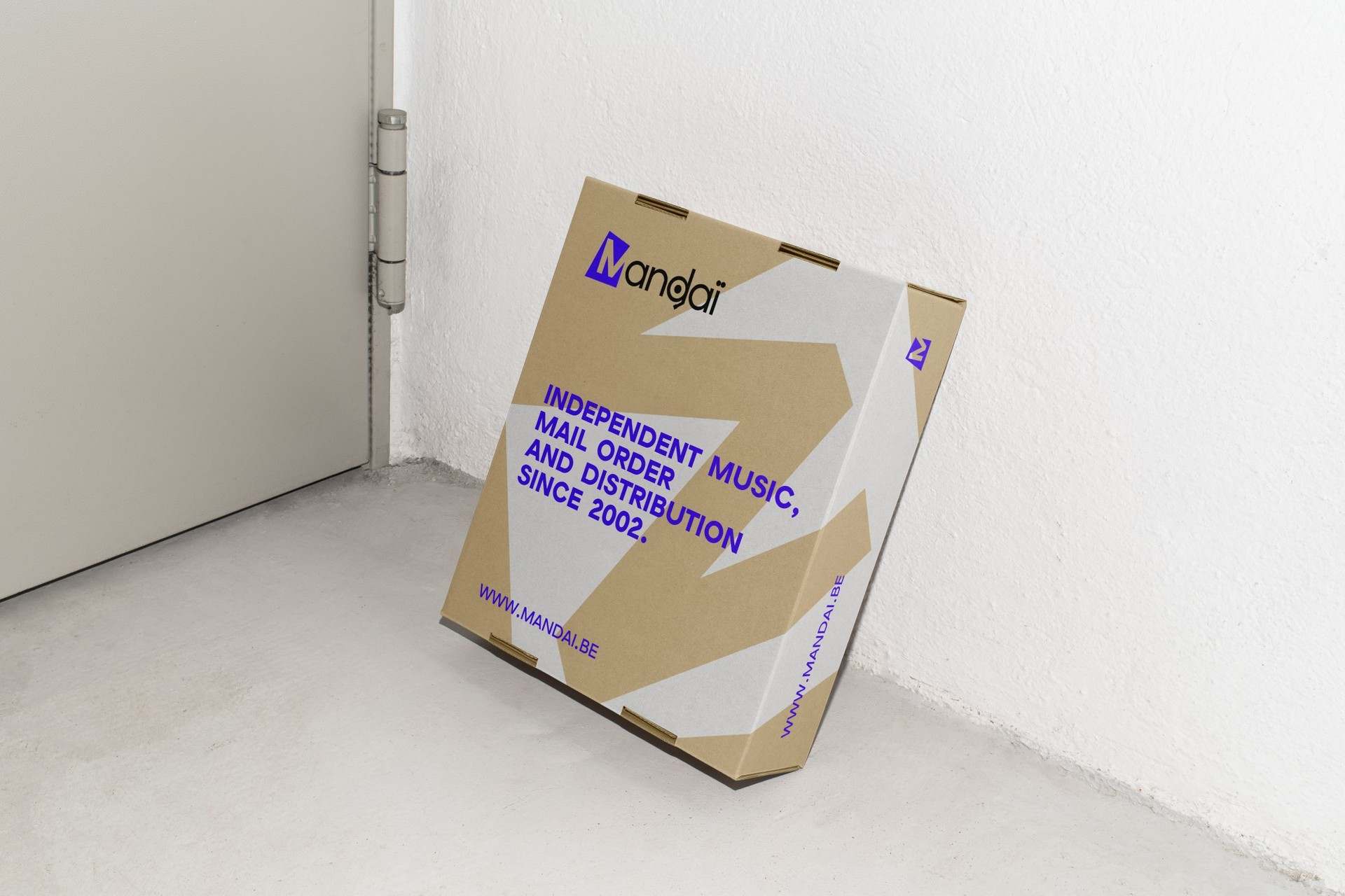 A kraft Mandaï shipping box leaning against a white wall, printed in violet with the shop's line and address.