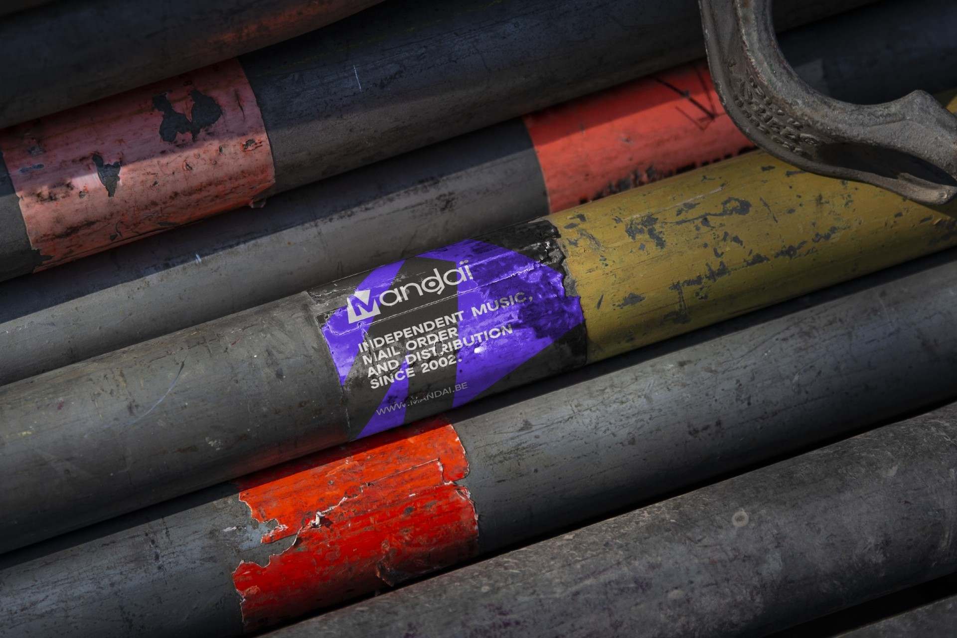 A black and violet Mandaï sticker wrapped around a worn scaffolding pole, among chipped red and yellow paint.